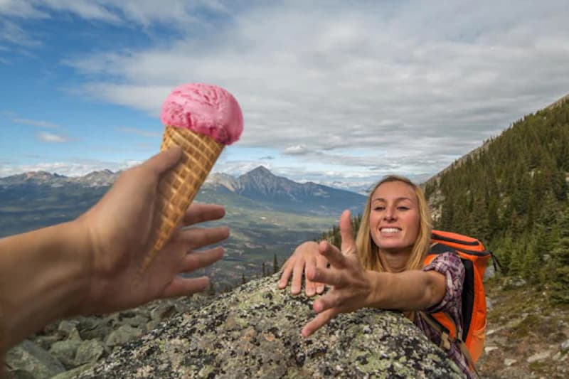 Frau mit Rucksack klettert über Fels und greift nach einer Eiswaffel, die von einer Hand entgegengehalten wird.