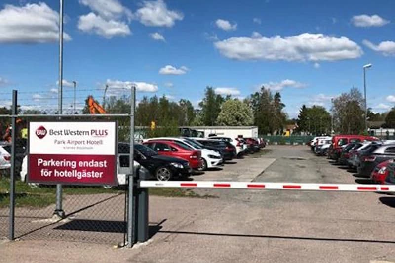 Geschlossener eingezäunter Parkplatz mit Schranke, Best Western PLUS Schild und geparkten Autos unter blauem Himmel.