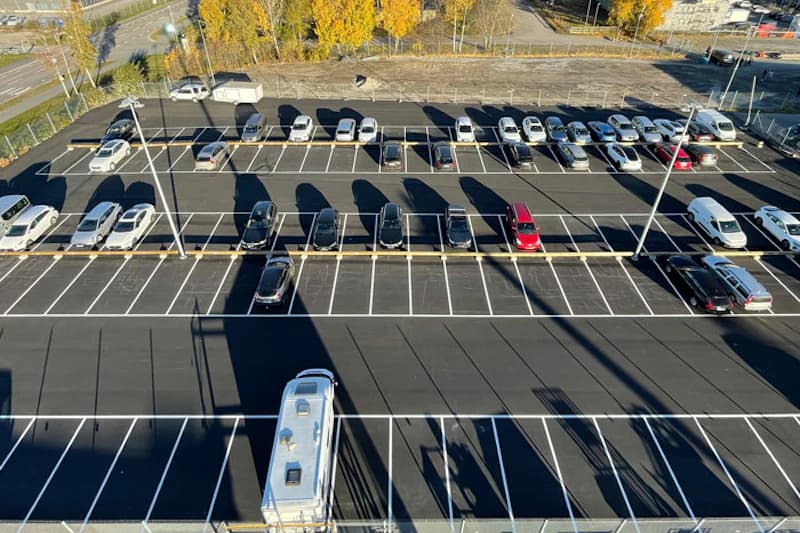 Vogelperspektive auf asphaltierten Parkplatz mit markierten Reihen, geparkten Autos und langen Lichtmastschatten.