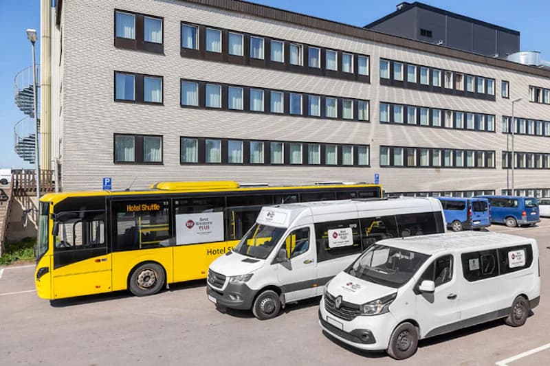 Gelber Hotel-Shuttlebus und zwei weiße Shuttle-Vans geparkt vor mehrstöckigem Gebäude mit Reihen von Fenstern.