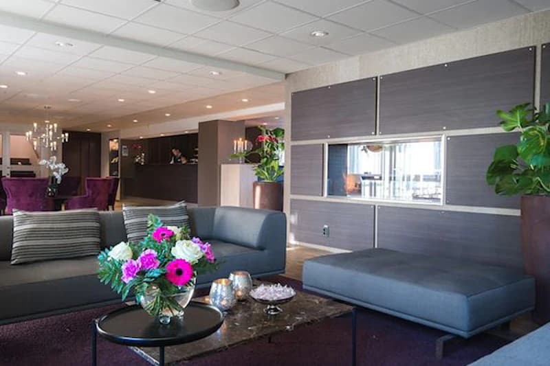 Moderne Hotellobby mit grauen Sofas, Blumenstrauß auf Tisch und Rezeption im Hintergrund.