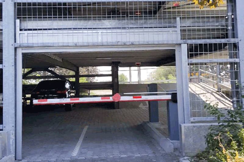 Einfahrt eines mehrgeschossigen Parkhauses mit abgesenkter Schranke, Metallgitter, Pkw im Inneren und Rampe rechts.