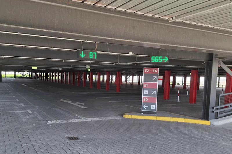 Überdachtes Parkhaus mit roten Stützen, grünen Leuchtdisplays und Richtungstafel für Parkebenen.