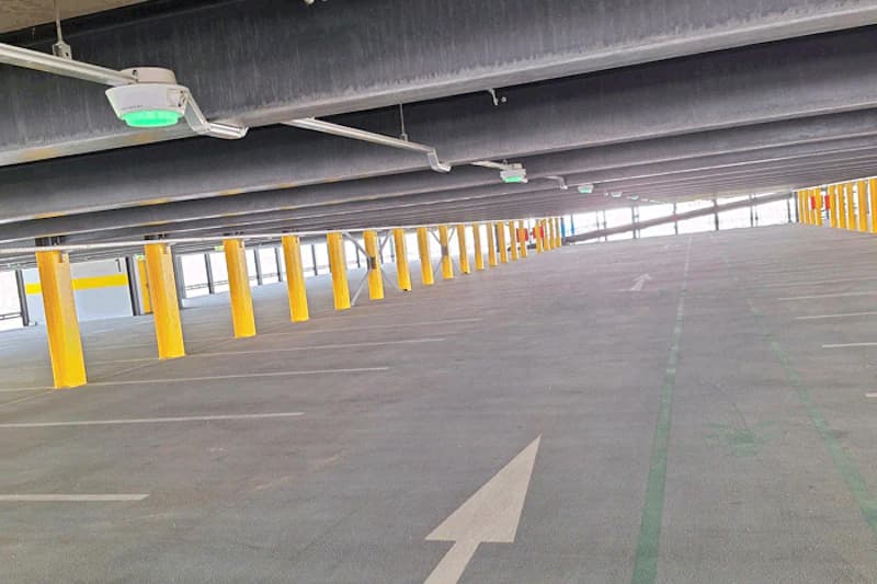 Leeres Parkdeck mit gelben Stützen, Deckenlampen mit grünen Lichtern und Pfeilen auf dem Boden.