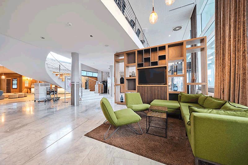 Weite Hotellobby mit grünem Sofa und Sesseln, Regalwand mit Fernseher und geschwungener Treppe.