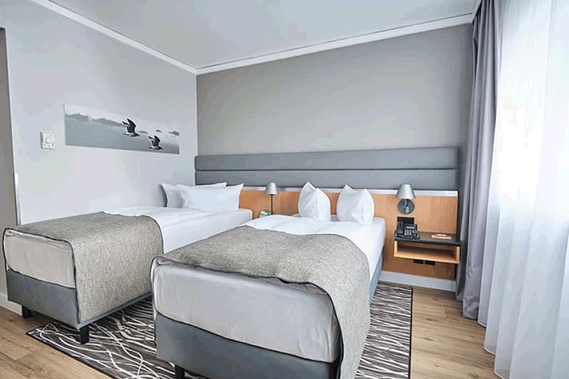 Zweibettzimmer mit zwei Einzelbetten, gepolstertem Kopfteil, Nachttisch mit Telefon, Tischlampen, Vorhang und Wandbild mit Vögeln.