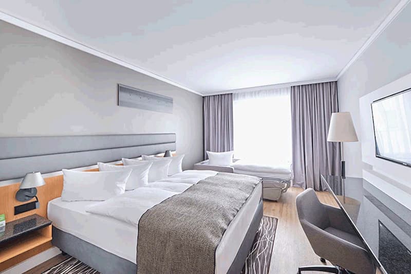 Helles Hotelzimmer mit großem Doppelbett, Tagesbett am Fenster, Schreibtisch, Stuhl und Wandfernseher.