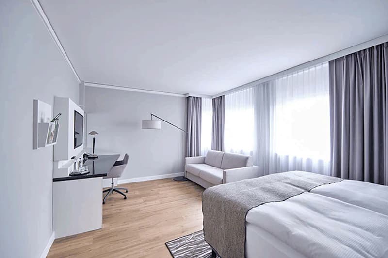 Helles Hotelzimmer mit Doppelbett, Sofa, Schreibtisch, Stehlampe und großen Fenstern mit Vorhängen.