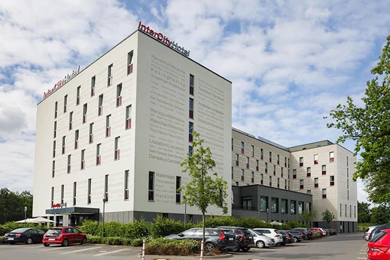 Mehrstöckiges weißes Hotelgebäude mit IntercityHotel-Schriftzug auf dem Dach, parkende Autos und Bäume im Vordergrund.