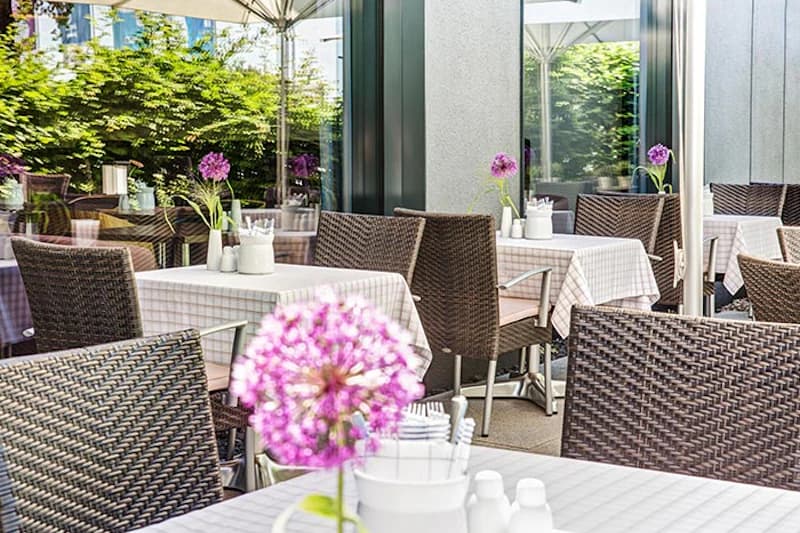 Freiluftterrasse mit gedeckten Tischen, geflochtenen Stühlen und lila Kugelblumen vor Glasfassade.