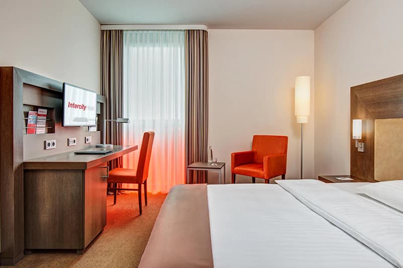Hotelzimmer mit Doppelbett, Schreibtisch mit TV (IntercityHotel), Fenster mit Vorhängen und zwei orangefarbene Sessel.