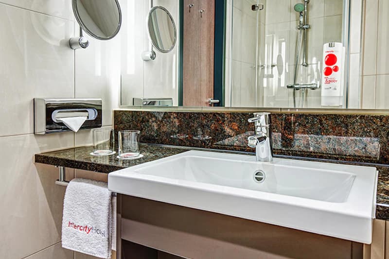 Weißes Waschbecken auf dunkler Granitabdeckung, Spiegel, zwei Gläser, Handtuch mit IntercityHotel-Logo und Duschkabine im Hintergrund.