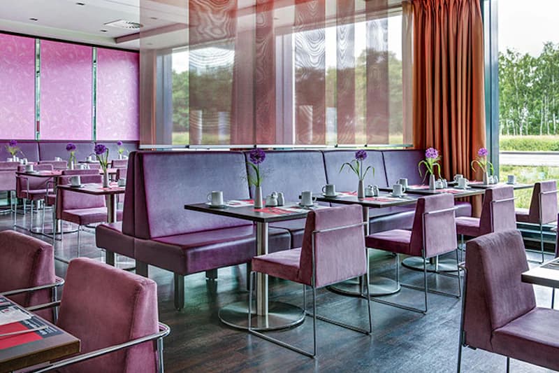 Speisesaal mit violetten Polstersesseln und -bänken, gedeckten Tischen und Blumen vor großen Fenstern.