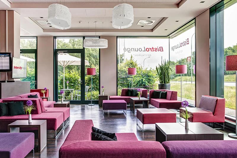 Hotel-Lounge mit pinken Sitzelementen, niedrigen Tischen, Steh- und Hängelampen und großen Glasfenstern zum begrünten Außenbereich.