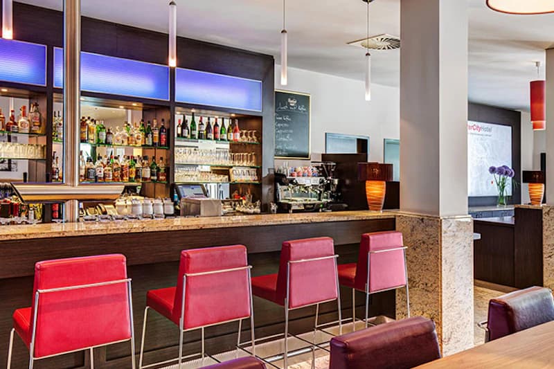 Hotelbar mit vier roten Barhockern, Marmortheke, beleuchteten Regalen mit Flaschen und Pendelleuchten.