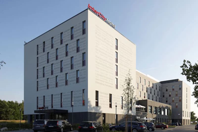 Weißes mehrstöckiges Hotelgebäude mit IntercityHotel-Schriftzug auf dem Dach und geparkten Autos im Vordergrund.