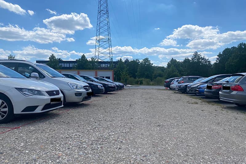 Schotterparkplatz mit geparkten Autos in Reihen, Strommast und Bäume unter blauem Himmel.