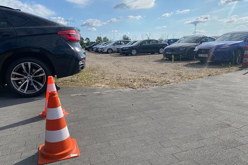 Pylone auf gepflasterter Fläche, dahinter geparkte Autos auf Schotter und ein dunkler SUV.