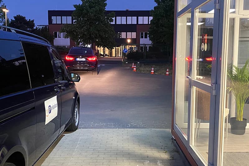 Dunkler Kleinbus in der Abenddämmerung vor einem beleuchteten Gebäude, daneben die offene Glastür des Empfangs.