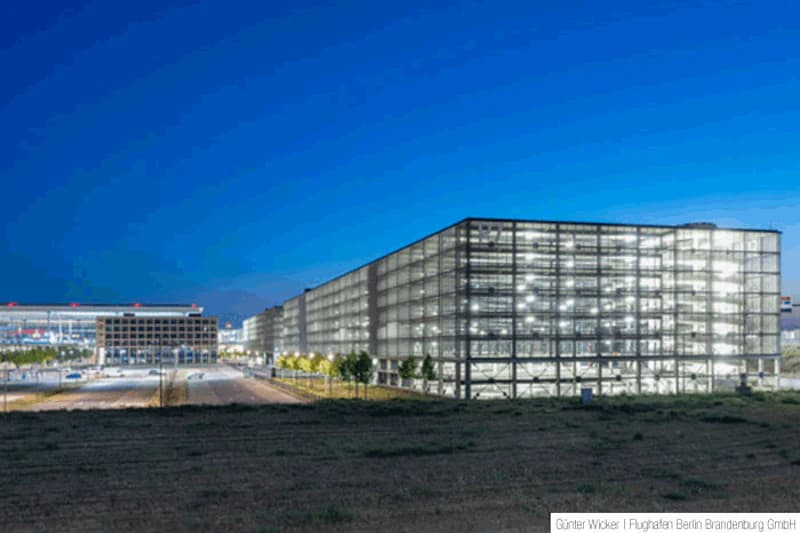 Beleuchtetes mehrgeschossiges Parkhaus mit Glasfassade neben Flughafen-Terminal bei Abenddämmerung.