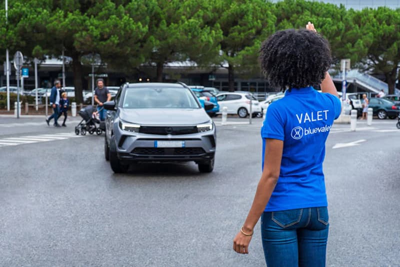 Parkservice-Mitarbeiterin in blauem Shirt weist ein graues Auto auf dem Flughafenparkplatz ein.