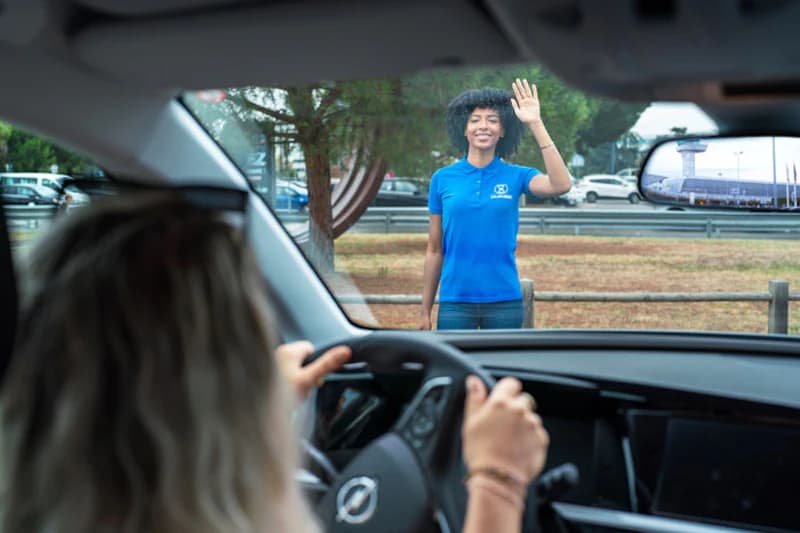 Blick aus dem Auto auf eine lächelnde Mitarbeiterin im blauen Poloshirt, die dem Fahrer zuwinkt.