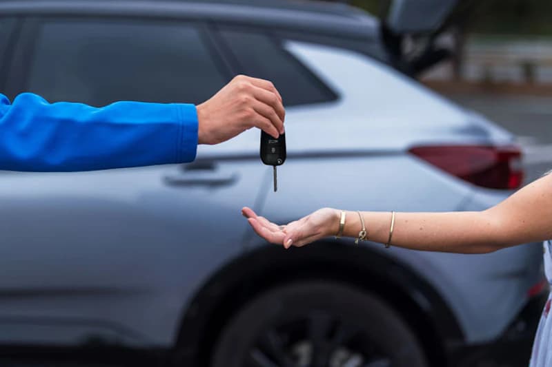 Hand übergibt Autoschlüssel an andere Hand vor grauem Auto.