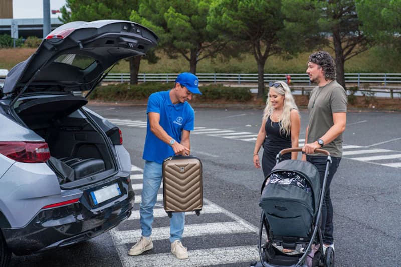 Parkmitarbeiter hebt Koffer aus geöffnetem Kofferraum vor wartendem Paar mit Kinderwagen auf Parkplatz.
