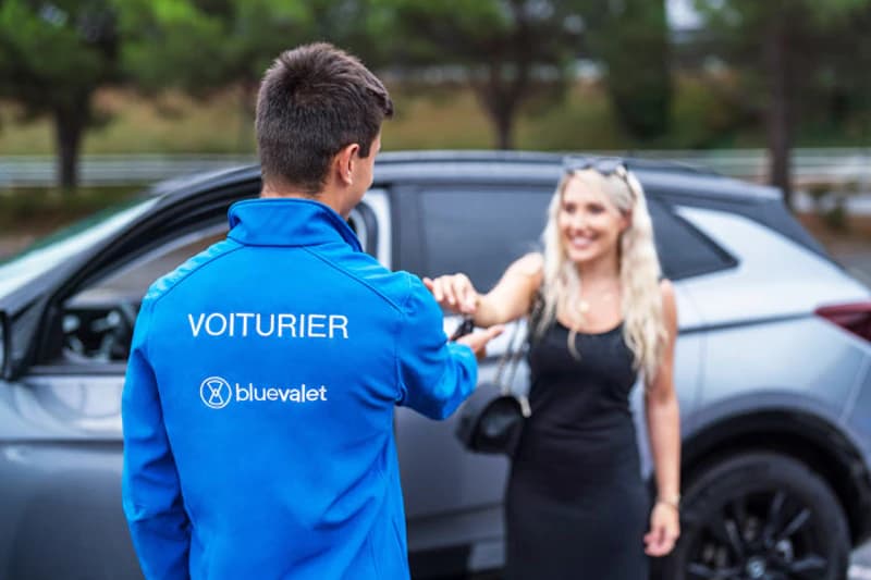 Parkmitarbeiter mit blauem Jackett VOITURIER übergibt Autoschlüssel an Frau vor silbernem SUV.
