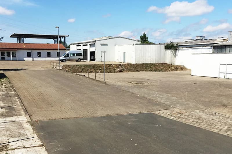 Leerer gepflasterter Parkplatz mit weißen Hallen, einem geparkten Kleinbus und einer Straßenlaterne unter blauem Himmel.