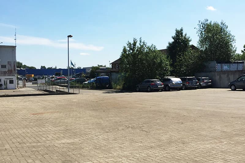 Großer gepflasterter Parkplatz mit vereinzelten geparkten Autos, Bäumen und Gebäuden im Hintergrund unter klarem Himmel.