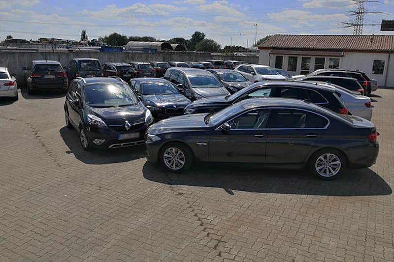 Parkplatz mit mehreren geparkten Autos auf gepflasterter Fläche und kleinem Bürogebäude.