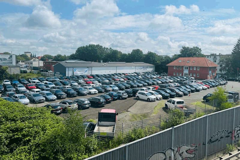 Großer umzäunter Parkplatz mit dicht gereihten Autos, Lagerhalle und rotem Gebäude im Hintergrund.