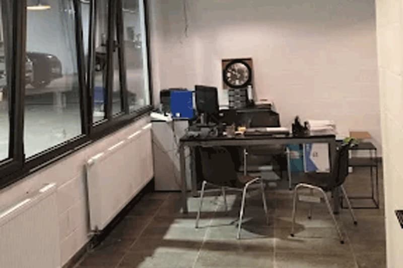 Kleines Büro mit Schreibtisch, zwei Stühlen, Computer und Wanduhr neben großem Fenster zum Parkhaus.