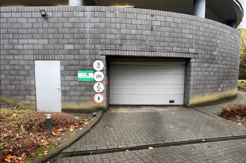 Geschlossene Parkhauseinfahrt mit Rolltor in grauer Klinkerwand, Nebentür und mehreren Verkehrsschildern am Portal.