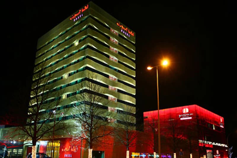 Mehrgeschossiges, nachts beleuchtetes Hotelgebäude mit roter Beschriftung Airport Hotel Basel und einem beleuchteten Nebengebäude.