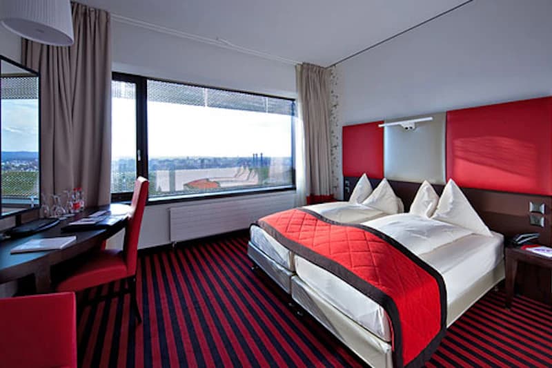 Zimmer mit zwei Betten, roten Kopfteilen, gestreiftem Teppich, Schreibtisch und großem Fenster mit Aussicht.