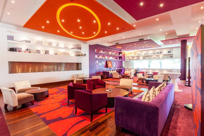Bunte Hotellounge mit violetten Sofas, roten Sesseln, gemustertem Teppich und Deckenbeleuchtung.