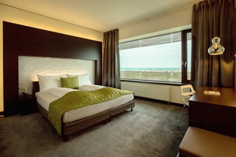 Hotelzimmer mit Doppelbett, grünem Überwurf, Kopfkissen, Schreibtisch und großem Fenster mit Außenblick.