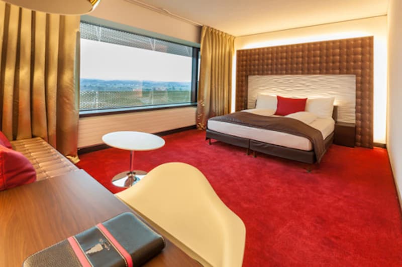 Hotelzimmer mit Doppelbett, tiefrotem Teppich, Schreibtisch, Sitzbank und breitem Fenster mit Außenblick.