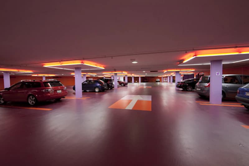 Blick in eine beleuchtete Parkgarage mit markierten Parkplätzen, Bodenpfeil und geparkten Autos.