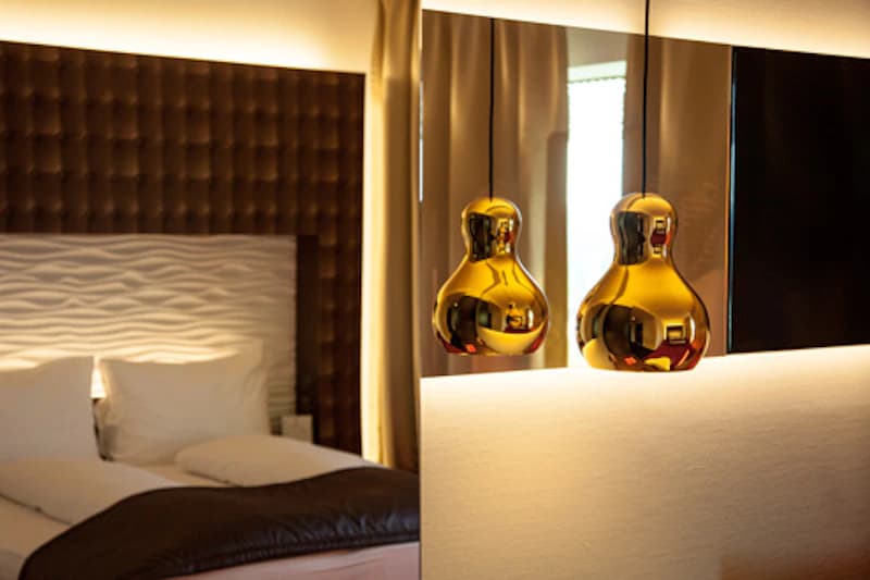 Hotelzimmer mit Bett, gepolstertem Kopfteil und goldenen Hängeleuchten neben einem Spiegel.