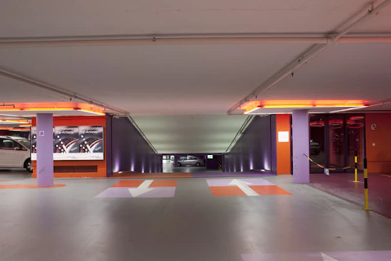 Parkebene mit Abfahrt zur unteren Etage, orange-violette Stützen, Bodenpfeile und geparkte Fahrzeuge im Hintergrund.