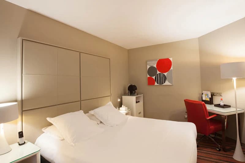 Hotelzimmer mit großem Doppelbett, gepolstertem Kopfteil, Nachttischen mit Lampen und Schreibtisch mit rotem Stuhl.