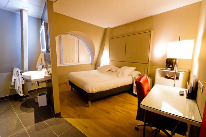 Schlafzimmer mit Doppelbett, Nachttischlampen, Schreibtisch mit rotem Stuhl und offenem Waschbereich mit rundem Fenster.