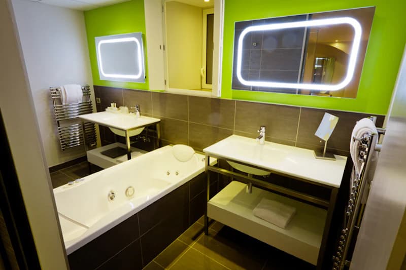 Modernes Badezimmer mit Badewanne, zwei Waschbecken, beleuchteten Spiegeln und grün gestrichener Wand.