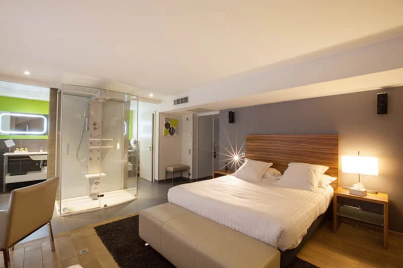 Doppelzimmer mit großem Bett, Holzkopfteile, offener Glasdusche, Nachttischlampen und Sitzbereich.