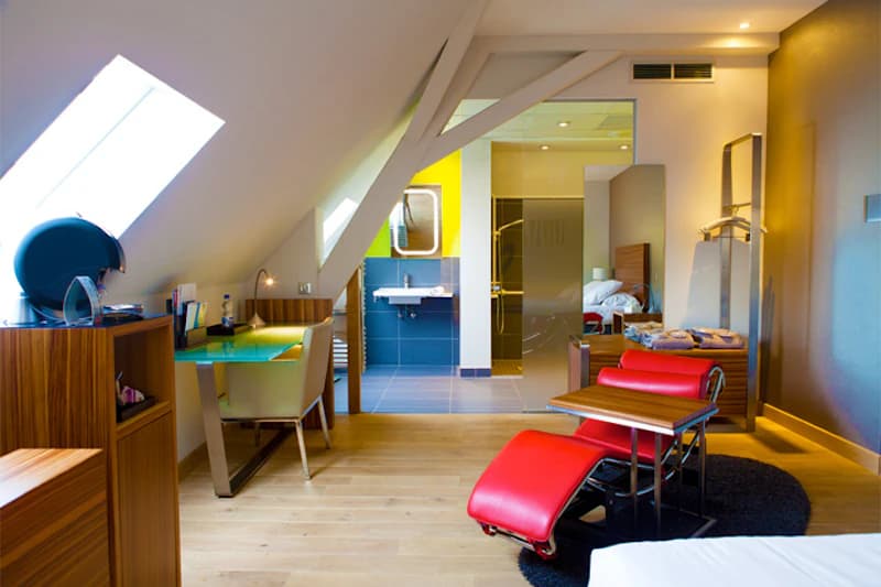 Dachzimmer mit roter Liege, Holzboden, Schreibtisch, Dachfenster und offenem Bad mit Waschbecken.