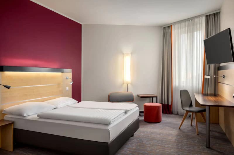 Vierstöckiges Hotel Vienna House Easy mit beleuchtetem Eingangsbereich, parkenden Autos und umzäuntem Gelände.