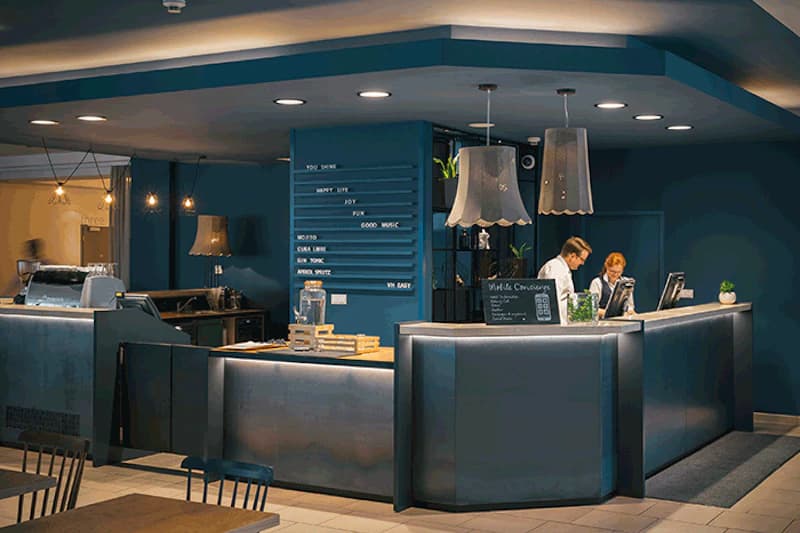 Rezeptionstheke in dunklem Petrol mit zwei Mitarbeitenden, Hängeleuchten und Menütafel im Hintergrund.